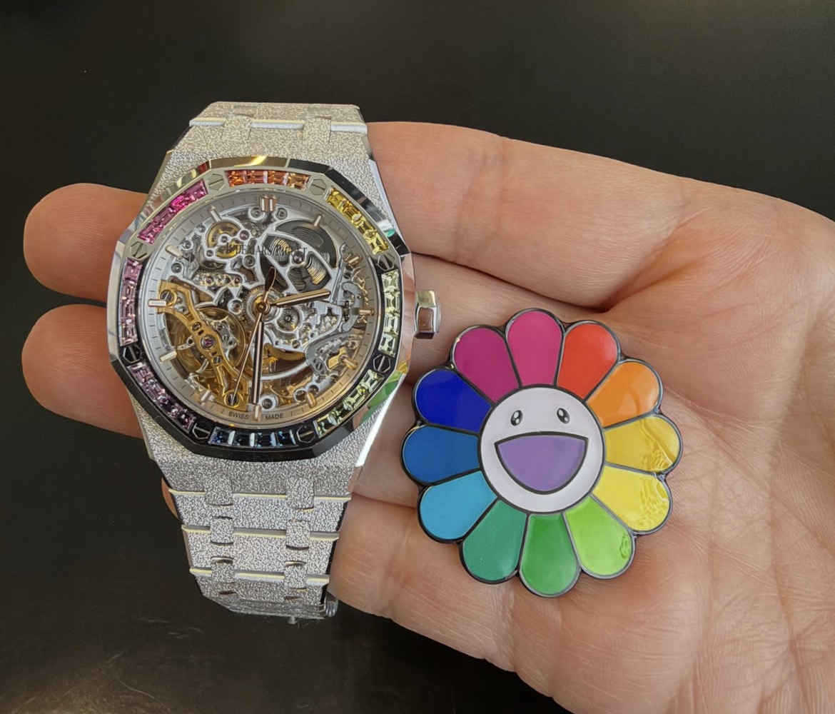 Audemars Piguet Royal Oak Double Balance Wheel Openworked 37mm in frosted white gold with a rainbow gem-set bezel, held beside a Takashi Murakami flower pin
